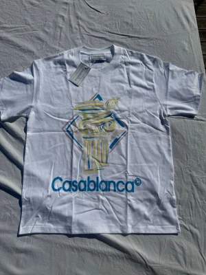Vit Casablanca t-shirt med tryck - Säljer en vit t-shirt från Casablanca med färgglatt tryck av en pelare och blå logga framtill. Klassisk passform och rund hals. Perfekt för dig som gillar stilrena plagg med en twist.