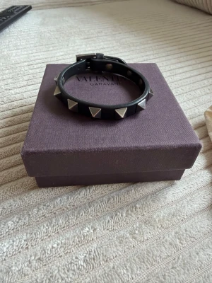 Valentino Garavani armband - Snyggt svart armband från Valentino i äkta läder med silvriga pyramidformade nitar runt om. Justerbart spänne. Kommer med original ask, påse och extra nitar. Perfekt accessoar för att lyfta din outfit. Släppt något i lädret, men det går att limma. Fint skick.