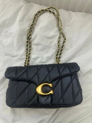 Svart quiltad handväska från Coach - Snygg svart quiltad handväska från Coach med guldfärgad kedjerem och stort C-spänne framtill. Väskan har innerfack med dragkedja och klassisk logga på insidan. Perfekt accessoar för att lyfta din outfit. Säljer den helt nytt efter som tyckte inte den passa mig skriv till mig om ni har fler frågor 
