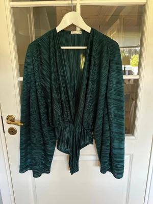 Mörkgrön plisserad body - Elegant body i en djup mörkgrön/teal nyans med vacker plisserad struktur. Modellen har en djup omlottringning, markerad midja och vida, böljande ärmar💫