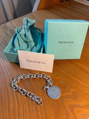 Tiffany & Co. silverarmband med berlock - Stilrent silverarmband från Tiffany & Co. med rund berlock där det står 'Please Return to Tiffany & Co. New York 925'. Armbandet har grova länkar och klassisk design. Levereras med original ask, påse och kort.