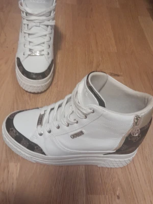 Vita och guldiga Guess sneakers strl 40 - Snygga sneakers från Guess i vitt konstläder med gulddetaljer och svarta partier med guldmönster. Skorna har dragkedja på sidan, högt skaft och tjock vit sula med mönstrad kant. Perfekta för dig som vill sticka ut med stilrena detaljer.