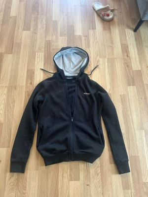 Svart hoodie från Mckenzie - Svart hoodie från McKenzie med dragkedja framtill och justerbar huva med grå insida. Klassisk passform med ribbade muddar vid ärmar och nederkant. Perfekt för chill dagar eller när du vill ha en enkel och stilren look.