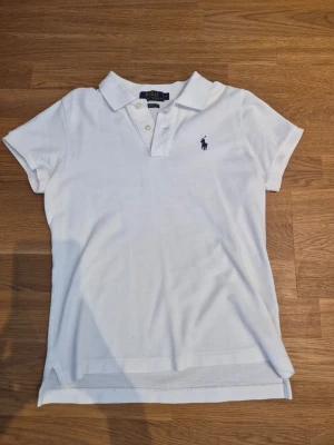 Vit pikétröja från Polo Ralph Lauren - Klassisk vit pikétröja från Polo Ralph Lauren med korta ärmar och mörkblå broderad logga på bröstet. Den är av 100% bomull.