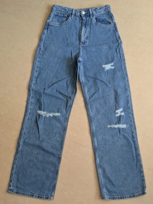 Blå raka jeans med slitningar - Säljer ett par blå raka jeans med trendiga slitningar på båda benen. Jeansen har hög midja, klassiska fem fickor och stängs med dragkedja och knapp. Perfekta för en avslappnad och cool stil.