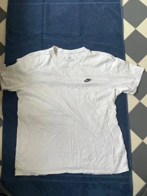 Vit t-shirt från Nike med logga - Klassisk vit t-shirt från Nike med liten svart logga på bröstet. T-shirten har rund halsringning och korta ärmar. Tillverkad i mjuk bomull för en skön känsla och enkel stil som funkar till det mesta.