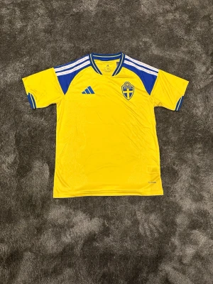 Svensk landslagströja - Gul kortärmad fotbollströja från Adidas med blå detaljer och vita ränder på axlarna. Bröstet har Sveriges landslagsemblem och Adidas-logga. Tillverkad i lätt och ventilerande material, perfekt för träning eller match.