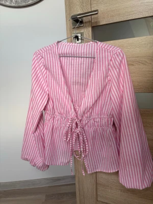Rosa och vitrandig blus med knyt - Säljer en söt blus med rosa och vita vertikala ränder. Blusen har djup v-ringning, långa ärmar och ett knytband i midjan för en snygg passform. Perfekt för en trendig och avslappnad look. Materialet känns lätt och luftigt.