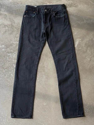 Levi’s 501 Straight Leg Jeans – Urtvättad Svart / Grå (L34) - Klassiska Levi's 501 med raka ben och knappgylf. Materialet är kraftig denim i en naturligt urtvättad svart/mörkgrå färg. Läderpatch baktill och klassisk Red Tab på bakfickan.
