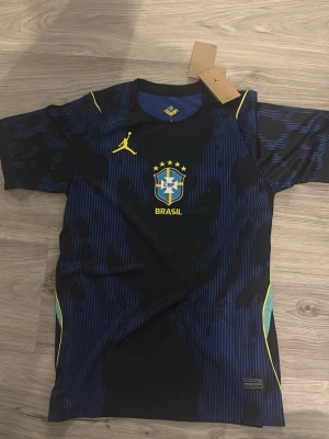 Brasil matchtröja Jordan x Nike - Snygg Brasil matchtröja i svart och mörkblått med gula detaljer. Tröjan har CBF-logga, fem stjärnor och BRASIL-tryck på bröstet samt Jordan-logga i gult. Tillverkad i lätt och ventilerande material, perfekt för fotboll eller träning storlek S, sitter som M.