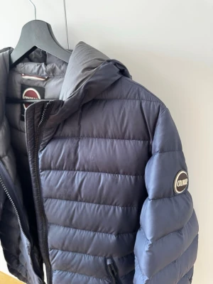 Mörkblå dunjacka från Colmar - Snygg mörkblå dunjacka från Colmar med huva och diskret logga på ärmen. Jackan har quiltad design, dragkedja framtill och två sidofickor med dragkedja.