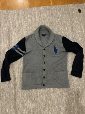 Ralph lauren cardigan - Snygg grå kofta med mörkblå ärmar från Polo Ralph Lauren. Stor blå logga på bröstet och nummerdetalj på ena ärmen. Koftan har sjalkrage, knäppning framtill och två fickor. Perfekt för dig som gillar sportig och klassisk stil.