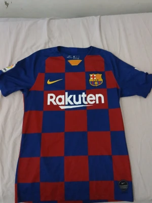 Barcelona matchtröja Nike röd/blå - Säljer en FC Barcelona matchtröja från Nike med klassiskt rutigt mönster i rött och blått. Tröjan har klubbmärke, Nike-logga och Rakuten-tryck på bröstet samt Dri-FIT-material som andas. Perfekt för dig som älskar fotboll och vill bära ditt lag.