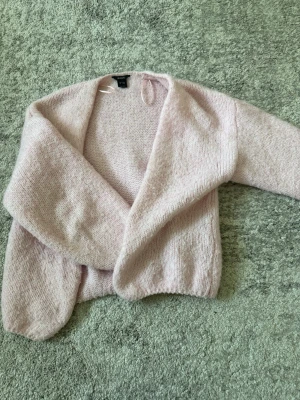 Ljusrosa stickad kofta från Lindex - Mysig ljusrosa stickad kofta från Lindex med öppen front och lång ärm. Koftan har en fluffig och mjuk känsla, perfekt att slänga över en topp. Enkel och stilren design som passar till många outfits.
