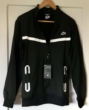 Svart zip hoodie plus byxor  från Nike - Svart hoodie och byxor från Nike med vit logga på bröstet och vita detaljer längs bröstet och vid fickorna. Dragkedja framtill och två fickor med dragkedja. Tillverkad i mjukt material som känns skönt mot huden. Perfekt för en sportig och avslappnad stil.