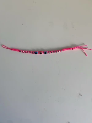 Rosa armband - Snyggt armband med rosa snöre, dekorerat med silverfärgade pärlor och tre ögonmotiv i mitten. Justerbar knytning för perfekt passform. 