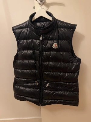 Svart dunväst från Moncler - Snygg svart dunväst från Moncler med hög krage och två dragkedjeförsedda fickor framtill. Västen har ett quiltat mönster och Moncler-logga på bröstet. Perfekt för lager-på-lager och ger en sportig vibe.
