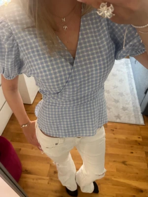 Blåvit rutig omlottopp - Supersöt blå och vit rutig omlottopp med korta puffärmar och markerad midja. Toppen har en v-ringad omlottdesign och ger en fräsch och somrig känsla. Perfekt att matcha med jeans eller kjol för en trendig look.