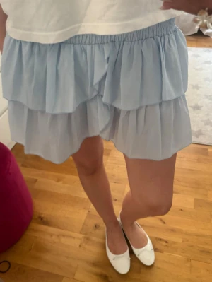 Ljusblå volangskort med resårmidja - Supersöta ljusblå skorts med dubbla volanger och elastisk midja. Luftigt och flowy material som ger en kjol-look men med shortsfunktion. Perfekt för en trendig och bekväm stil. Märke och material är ej angivet.