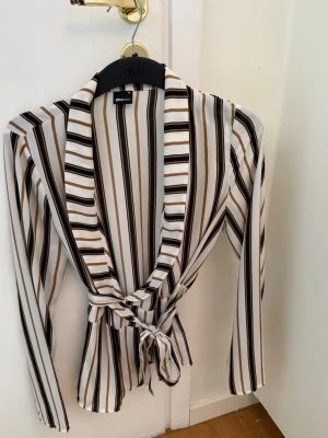 Randig blus med knytband från Gina - Snygg blus från Gina med vertikala ränder i vitt, svart och brunt. Blusen har långa ärmar, djup urringning och ett knytband i midjan som ger en feminin siluett. Perfekt för att lyfta vilken outfit som helst.