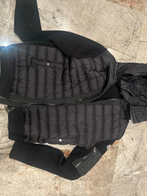 Moncler cardigan - Snygg svart pufferjacka från Moncler med quiltad kropp och stickade ärmar samt huva. Jackan har dragkedja framtill, fickor med knapp och en Moncler-logga på ärmen. Perfekt för kyliga dagar och ger en sportig men stilren look.