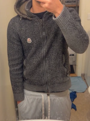 Grå ziptröja med huva från Moncler - Säljer en grå stickad ziptröja från Moncler med huva och ribbade muddar. Tröjan har ett Moncler-märke på bröstet och två fickor med dragkedja. Perfekt för dig som gillar stilrena och varma plagg.