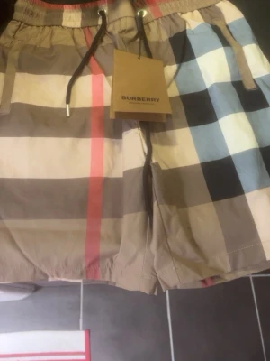 Rutig Burberry shorts med snörning - Säljer ett par klassiska Burberry shorts med det ikoniska rutiga mönstret i beige, svart, rött och blått. Shortsen har elastisk midja med snörning och är tillverkade i ett lätt och bekvämt material, perfekt för sommaren.