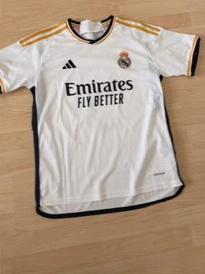 Real Madrid matchtröja Adidas S - Säljer en vit Real Madrid matchtröja från Adidas i storlek S. Tröjan har guldfärgade detaljer på axlarna, svart och gul krage samt klubbmärke och Adidas-logga på bröstet. Tillverkad i lätt och ventilerande material, perfekt för fotbollsträning eller match.