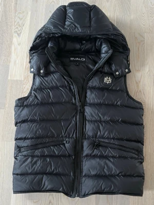 Bvaldi väst - Snygg svart dunväst från Bvaldi med avtagbar huva och två dragkedjefickor framtill. Västen har ett stilrent emblem på bröstet och quiltad design som ger en modern look. Perfekt för lager-på-lager och streetwear-vibe. Storlek S, Bra skick