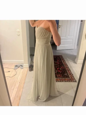 Ljus axelbandslös långklänning/ med band runt axlarna - Elegant axelbandslös långklänning i ljus beige ton med vackert draperad överdel och markerad midja. Klänningen har ett flytande fall och är tillverkad i ett tunt, mjukt material som ger en romantisk känsla. Perfekt för speciella tillfällen.