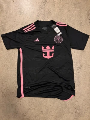 Svart Inter Miami matchtröja Adidas (S/M) - Snygg svart matchtröja från Inter Miami CF med rosa detaljer och logga på bröstet. Tröjan har korta ärmar, rund hals och tre rosa ränder på axlarna. Tillverkad i lätt och ventilerande material med Aeroready-teknologi. (Stor sitter som M)