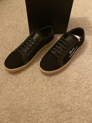 Svarta Saint Laurent sneakers - Snygga svarta sneakers från Saint Laurent med vit text på sidan och beige sula. Skorna har en klassisk låg profil, svarta skosnören och är tillverkade i en mix av läder och textil. Perfekta för dig som vill ha en clean och stilren look.