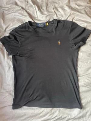 Svart t-shirt från Polo Ralph Lauren - Klassisk svart t-shirt från Polo Ralph Lauren med broderad logga på bröstet. Modellen är custom slim fit och har korta ärmar samt rund halsringning. Tillverkad i mjuk bomull för en skön känsla och stilren look. MODELLEN ÄR 181CM