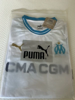 Marseille matchtröja Puma vit/blå - Säljer en vit Marseille matchtröja från Puma med blå detaljer och klubbmärke på bröstet. Tröjan har rund hals med blå kant, guld Puma-logga och sponsortryck framtill. Tillverkad i lätt och ventilerande material, perfekt för fotbollsträning eller match.