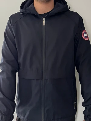 Svart vindjacka från Canada Goose - Snygg svart vindjacka från Canada Goose med huva och dragkedja framtill. Jackan har två sidofickor med dragkedja och den klassiska Canada Goose-loggan på vänster ärm. Perfekt för blåsiga dagar och enkel att matcha med olika outfits. Storleken är XL men passform är Medium. HELT NY SKICK.