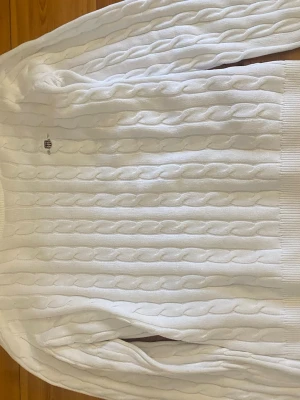 Vit kabelstickad tröja Gant - Kabelstickad vit tröja från Gant med rund halsringning och diskret broderad logotyp på bröstet. Tröjan har ribbade muddar vid ärmslut och nederkant, och är tillverkad i mjukt material som ger en klassisk och stilren look.