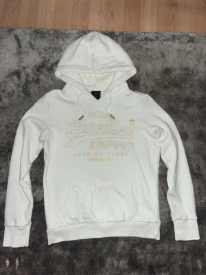 Vit Superdry hoodie med gulddetaljer - Säljer en vit hoodie från Superdry med guldfärgat tryck och japanska tecken på bröstet. Hoodien har en klassisk huva med dragsko, magficka och ribbade muddar vid ärmar och nederkant. Perfekt för dig som gillar streetwear och snygga detaljer. Hoodien är storlek XS