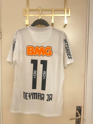 Vit Santos Neymar Jr matchtröja - Säljer en vit Santos FC matchtröja med Neymar Jr och nummer 11 på ryggen. Tröjan har tryckta sponsorer som BMG och Netshoes, samt Santos klubbmärke på bröstet. Tillverkad i lätt och ventilerande material, perfekt för fotboll eller samling.