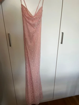 Ljusrosa långklänning med spetsdetaljer - Säljer en lång, ljusrosa klänning med tunna axelband och cowl neck. Klänningen är täckt av ett spetslager och har en elegant, figurnära passform. Storlek XS. Säljer då den inte kommer till användning. Aldrig använd, prislappen kvar.
