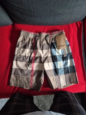 Rutig Burberry shorts  - Säljer ett par klassiska Burberry shorts med ikoniskt rutigt mönster i beige, svart, vit och röd. Shortsen har elastisk midja med svart snörning och sidofickor. Perfekta för sommaren och har en avslappnad passform. De är M men de passar S bäst eftersom på M ÄR den tight