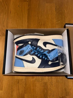 Nike Air Jordan 1 High Obsidian blå/vit - Säljer ett par Nike Air Jordan 1 High ”Obsidian” sneakers i blått, vitt och marinblått läder i storlek 41. Klassisk siluett med högt skaft, perforerad tåbox och ikoniska Swoosh-loggan på sidan. Skorna har blå skosnören och extra svarta snören medföljer.