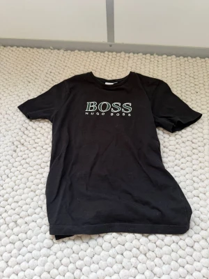 Svart Hugo Boss t-shirt - Snygg svart t-shirt från Hugo Boss med klassisk logga tryckt i vitt och grönt på bröstet. T-shirten har rund halsringning och korta ärmar. Perfekt basplagg för en stilren och enkel look.