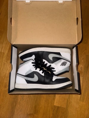 Svarta och vita Air Jordan 1 High - Säljer ett par klassiska Air Jordan 1 High sneakers i svart och vitt med grå swoosh i storlek 42. Skorna har högt skaft, svart snörning och ikoniska Jordan-loggan på sidan. Tillverkade i läder och textil för en clean och tidlös look.