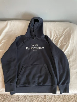 Peak Performance hoodie - (Pris kan diskuteras) Snygg mörkblå hoodie från Peak Performance med stor vit logga och text 'Est. 1986' på bröstet. Klassisk känguruficka framtill, ribbade muddar och justerbar huva med dragsko. Perfekt för en avslappnad och sportig stil.