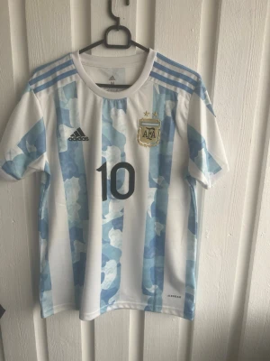 Argentina Messi matchtröja Adidas - Säljer en officiell Argentina fotbollströja från Adidas med Messi och nummer 10 på ryggen. Tröjan har klassiska blå och vita ränder, broderad AFA-logga, svarta Adidas-detaljer och Copa America-märke på ärmen. Tillverkad i lätt och ventilerande material.