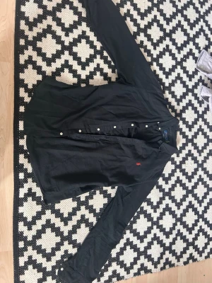 Svart skjorta från Ralph Lauren - Svart långärmad skjorta från Ralph Lauren med klassisk krage och knappar framtill. Skjortan har en röd broderad logotyp på bröstet och är tillverkad i bomull. Perfekt för en stilren och avslappnad look.