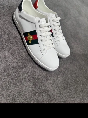 Vita Gucci sneakers med bi-detalj - Snygga vita sneakers från Gucci med klassiska gröna och röda ränder på sidan samt en guldfärgad bi-brodyr. Skorna har vita snören och detaljer i rött och svart på hälen. Tillverkade i slätt läder för en clean look. Man får med box och Gucci påse 
