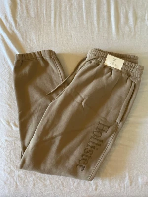 Beige mjukisbyxor från Hollister - Snygga beige mjukisbyxor från Hollister med broderad logga längs benet. Byxorna har dragsko i midjan och ribbade muddar vid bensluten. Tillverkade i mjukt och skönt material, perfekta för chill dagar. Klassisk passform och fickor på sidorna.