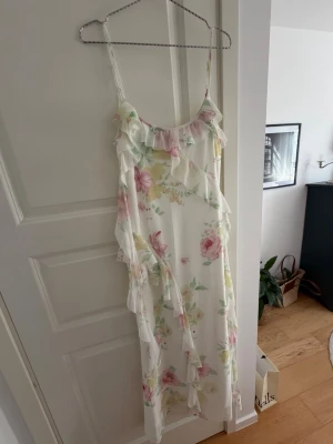 Blommig långklänning med volanger - Romantisk långklänning med tunna axelband och volangdetaljer. Klänningen är vit med blommigt mönster i rosa, gul och grön. Perfekt för sommarens alla tillfällen. Materialet känns lätt och luftigt, troligtvis polyester eller liknande.