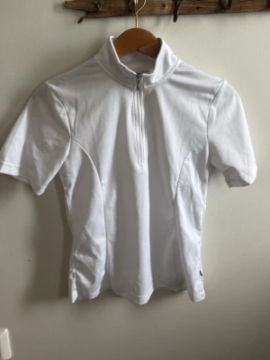 Vit polotopp med half zip - Snygg vit polotopp med kort ärm och half zip framtill. Tillverkad i ett stretchigt syntetmaterial som ger en sportig och clean look. Perfekt till dig som gillar minimalistisk stil och vill ha något bekvämt och stilrent. Tävligströja, använd 1 gång